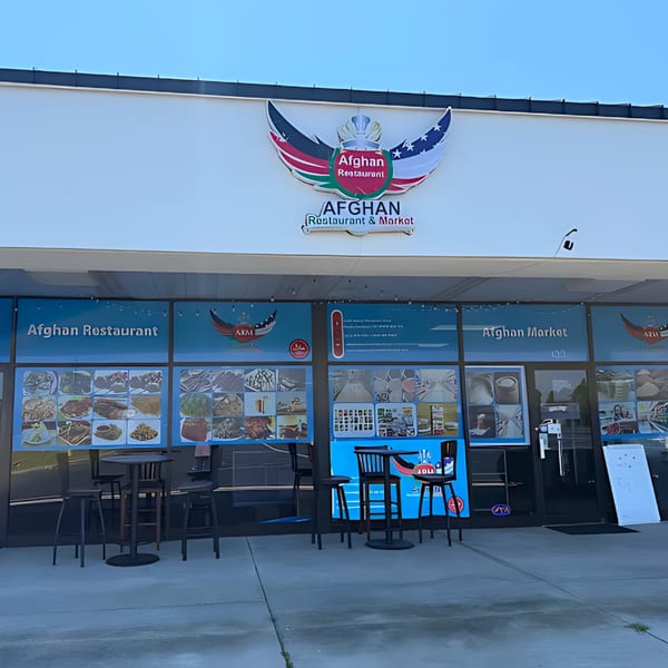 Exterior of Afghan Restaurant and Market with a winged logo, window displays of food, and outdoor seating.